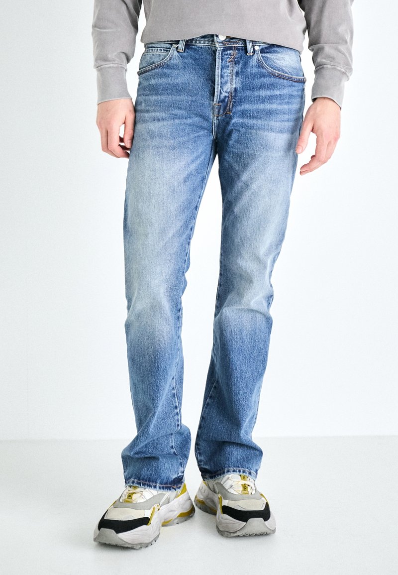 Vaqueros de mezclilla azul claro con corte recto, acabado desgastado y costuras en contraste, combinados con zapatillas gruesas en gris y amarillo.