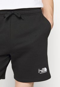 Short noir à cordon avec logo et slogan The North Face "Never Stop Exploring" sur la partie inférieure de la jambe gauche, porté par une personne avec la main dans la poche.