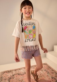 Horgolt rojtos póló, színes grafikai mintával a Rolling Stones logóival, csíkos farmer rövidnadrággal párosítva. Világos színek, hétköznapi stílus.