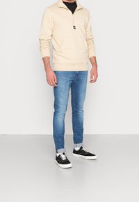 Felpa beige con zip e tasca frontale, abbinata a jeans blu e sneakers nere. La felpa ha una finitura testurizzata e polsini a costine.