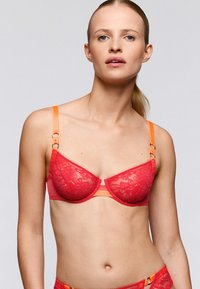 Soutien-gorge en dentelle rouge avec bretelles orange, présentant un design classique, des motifs floraux et des accents en métal doré. Texture lisse et forme maintien.