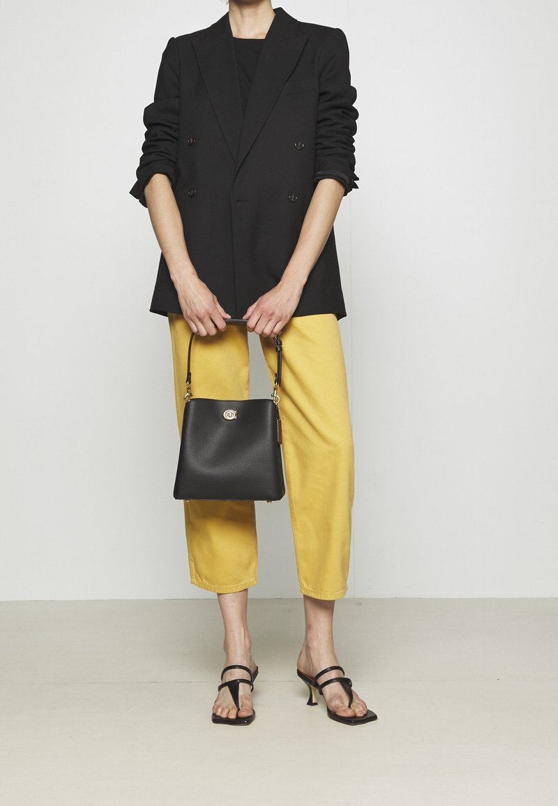 Personne portant un blazer noir et un pantalon court jaune tenant un sac à main noir, debout avec des sandales à talons noires à brides.