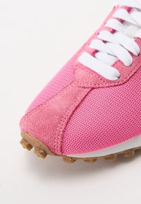 Gros plan d'une basket rose avec des lacets blancs, une tige en daim et en mesh, et une semelle en caoutchouc marron avec des crampons texturés.