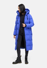 Lang pufferjas in helderblauw, met een capuchon en zwarte rits. Gecombineerd met een zwarte skinny jeans en zwarte veterschoenen. Gladde textuur.