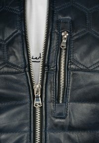 Marine lederen leren jas met een gebogen hexagonaal patroon, dubbele zilveren ritsen en een witte stof die eronder zichtbaar is. Textuur is duidelijk zichtbaar.