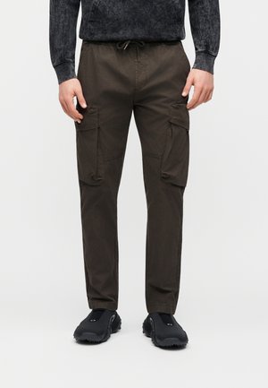 Hombre con camiseta de manga larga gris oscuro y pantalones cargo marrones con grandes bolsillos laterales, combinados con zapatos negros sin cordones.