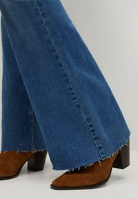 Jean en denim bleu avec ourlets bruts porté avec des bottines marron en daim à talons épais sur un fond uni.