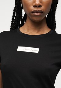 Κοντινό πλάνο ενός ατόμου που φοράει μαύρο T-shirt με μια μικρή λευκή ορθογώνια ετικέτα στο στήθος που γράφει "Armani Exchange".