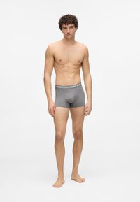 Jaunas vyras stovi basomis, vilkėdamas pilkus boxer trumpikes su prekės ženklo juosta, neutralia išraiška, vienodame šviesiame fone.