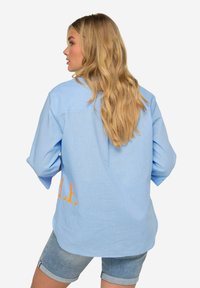 Jasnoniebieska koszula z długim rękawem w luźnym kroju, z pomarańczową grafiką „LE” po lewej stronie i podwiniętymi rękawami. Widoczne dżinsowe szorty.