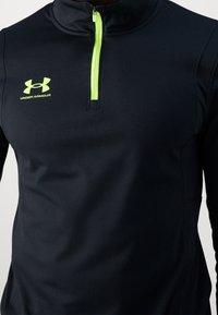 Svart idrottströja med halv dragkedja, med en neongrön dragkedja och logotyp, tillverkad av ett slätt, elastiskt material.