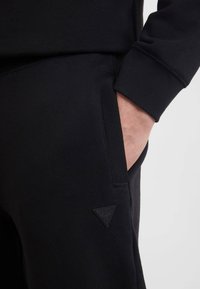 Schwarze Sweatpants mit glatter Textur, ausgestattet mit einer sichtbaren Seitentasche und einem kleinen schwarzen Dreieckslogo am unteren Bein.