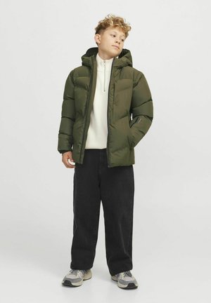 Giacca puffer verde scuro con cappuccio, chiusura con zip e tasche laterali. Indossata sopra un top bianco con collo alto e pantaloni neri a gamba larga. Tessuto strutturato.