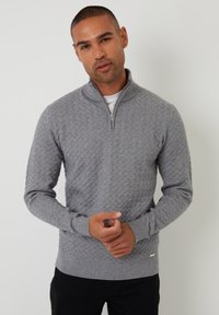 Šedý svetr na zip s texturovaným vzorem, dlouhými ruk�ávy a žebrovanými manžetami. Stylizovaný přes bílou košili, s logem na spodním lemu.