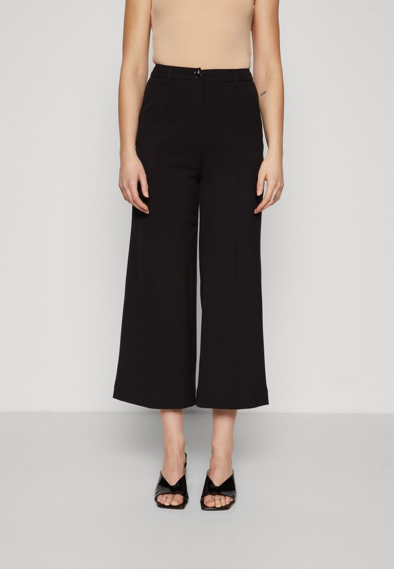 Pantalon noir à jambes larges avec une taille haute, doté d'une fermeture avant par bouton et fermeture éclair, en tissu lisse. Associé à des chaussures noires ouvertes à l'avant.