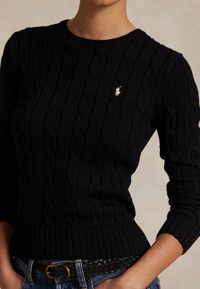 Primo piano di una persona che indossa un maglione nero a trecce con un piccolo logo bianco Polo Ralph Lauren sul petto.