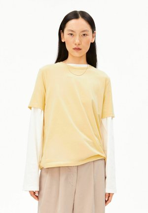 Vrouw met lang donker haar draagt een lichtgeel kort mouw topje over een wit hemd met lange mouwen en losse beige broek.