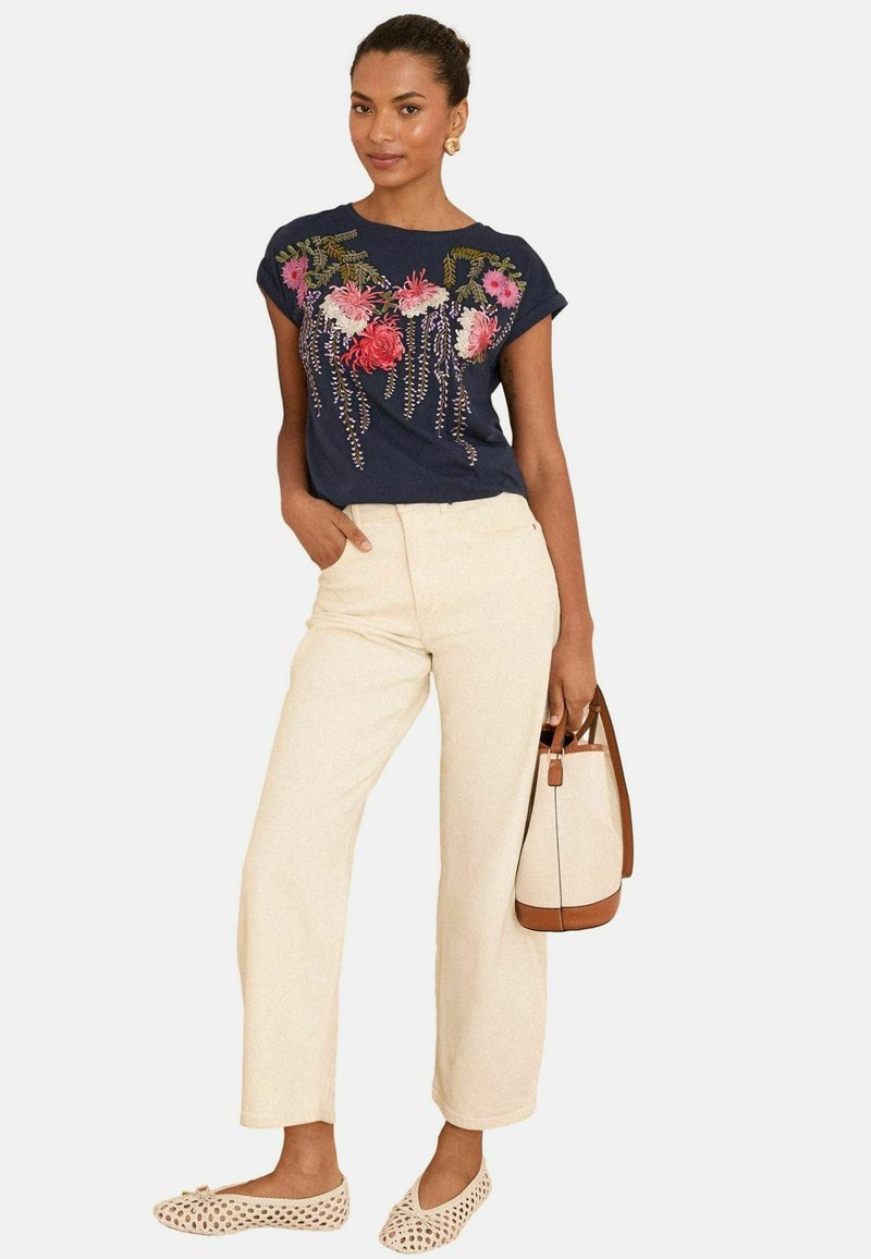 T-shirt navy a maniche corte con ricami floreali, abbinato a pantaloni larghi color crema e scarpe basse perforate di un chiaro color cammello. Borsa marrone in abbinamento.