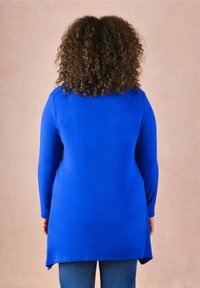 Långärmad topp i ljusblått tyg, med rundad nederkant och en avslappnad passform, sedd bakifrån mot en enfärgad bakgrund.