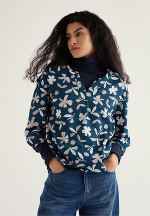 Vrouw met lang, donker krullend haar, gekleed in een marineblauw coltrui onder een blouse met een blauw bloemenpatroon en blauwe spijkerbroek, met haar handen in haar zakken.