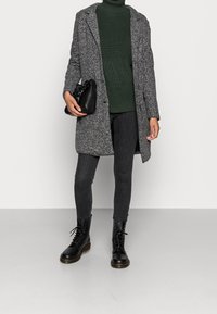 Abrigo texturizado gris sobre un suéter de cuello alto de canalé verde oscuro, combinado con jeans skinny negros y botas de combate con cordones. Bolso negro con un pequeño asa.
