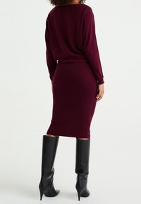 Burgunderröd stickad klänning med en figurnära passform och ribbad textur, i kombination med höga, blanka svarta knähöga stilettklackar.