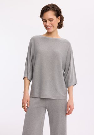 Uśmiechnięta kobieta w luźnym, jasnoszarym dzianinowym topie z rękawami do łokcia oraz pasujących spodniach, stojąca na jednolitym tle.