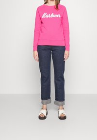 Sweat-shirt rose avec le logo "Barbour" blanc, associé à un jean en denim foncé et des sandales blanches à brides croisées. Design simple et coupe décontractée.