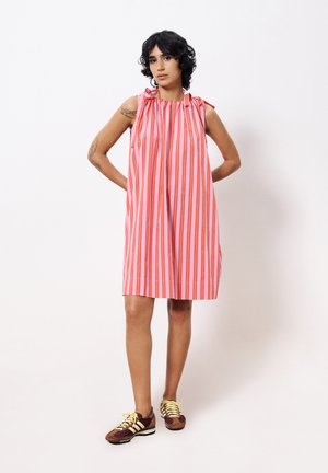 Robe sans manches rose avec des rayures rouges verticales, décolleté froncé et poches latérales. Associée à des baskets marron avec des accents jaunes.