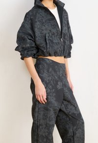 Veste noire raccourcie et pantalon assorti, avec un motif floral, réalisés dans un tissu texturé à finition légèrement brillante.