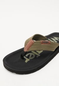Sandale à brides tressées vert olive avec accent en cuir marron. Semelle noire texturée comportant le logo "Wrangler" en jaune vif.
