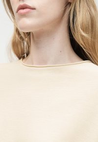 Jeune femme à la peau claire et aux cheveux blonds portant un haut côtelé crème, montrant le cou et le bas du visage sur un fond clair.