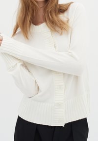 Krémový kardigán z měkkého materiálu, se žebrovanými rukávy a dole, s zapínáním na knoflíky a texturovanými akcenty uprostřed.