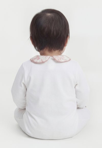 Baby med kort mørkt hår som sitter vendt bort, iført et hvitt antrekk med en rosa blomstret Peter Pan-krage.