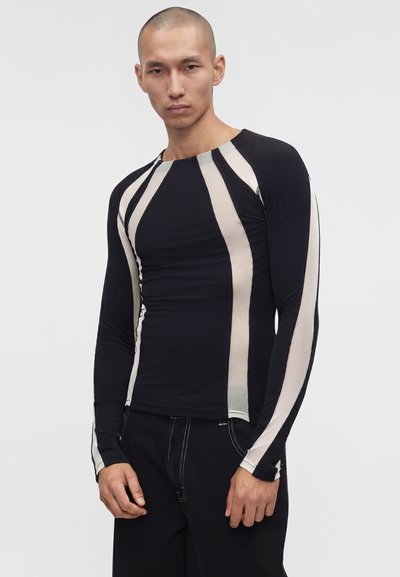 Άνδρας που φοράει εφαρμοστό μαύρο μακρυμάνικο πουκάμισο με κατακόρυφες μπεζ ρίγες και μαύρο παντελόνι, σε απλό φωτεινό φόντο.