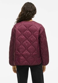 Femeie cu păr lung și creț, purtând o jachetă matlasată de culoare violet închis, cu cusături în formă de diamant, asortată cu pantaloni negri, văzută din spate.