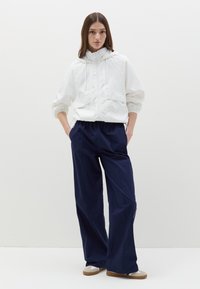 Giacca leggera bianca con cappuccio e polsini elasticizzati, abbinata a pantaloni blu navy a vestibilità ampia con vita elastica e tasche laterali.