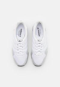 Pár bílých tenisek Adidas se šedými nubukovými špičkami, černými podrážkami a tkaničkami, z pohledu shora na bílém pozadí.