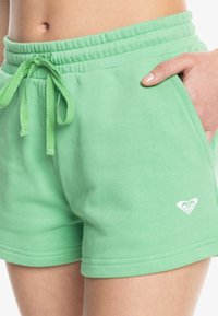 Shorts de sport en coton vert avec une taille élastique, un cordon de serrage ajustable, des poches latérales et un petit logo blanc sur l'ourlet.