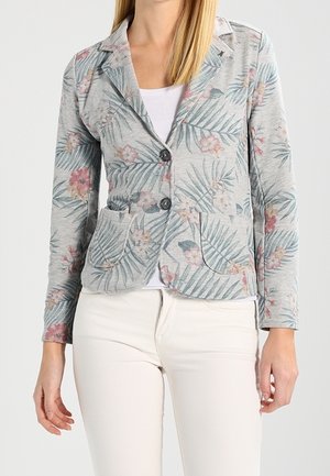 Un modèle porte un blazer gris clair avec un imprimé floral rose et feuilles vertes, sur un haut blanc et un pantalon crème.