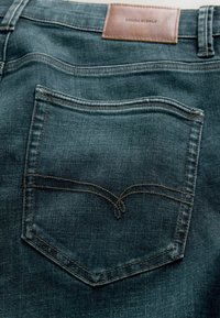 Tumed denim teksad, mille taga on tasku, millel on kaardus õmblus. Vöökoha peal on pruun nahk silt, millel on kirjas "DENIM SUPPLY". Sile tekstuur.