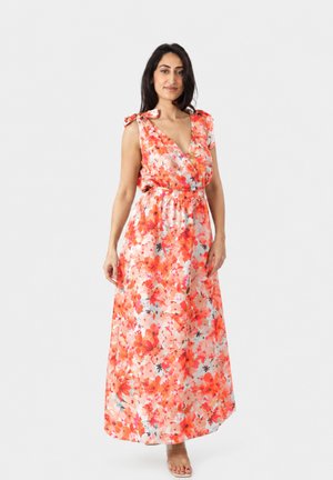 Donna che indossa un abito lungo senza maniche con stampa floreale arancione, rossa e rosa, scollo a V e spalline annodate, in piedi davanti a uno sfondo semplice.