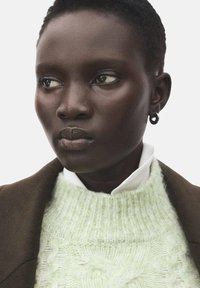 Pletena džemper u pastelnom zelenom s teksturiranim uzorcima, slojevito preko bijele košulje s ovratnikom, uparen s smeđom jaknom, s naušnicom u obliku obruča.