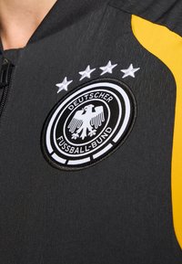 Sort sportsjakke med rund, brodert Deutscher Fußball-Bund-logo med en ørn, fire hvite stjerner og en gul aksentpanel.