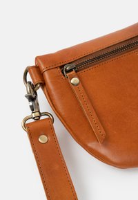 Braune Leder-Crossbody-Tasche mit runder Form, einer vorderen Reißverschlusstasche und einem abnehmbaren Riemen mit metallischen Hardware-Akzenten.