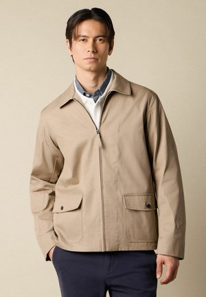 Młody mężczyzna w beżowej kurtce na suwak z patkami na kieszeniach, nałożonej na białą koszulę i ciemnoniebieskie spodnie, stojący na jednolitym tle.