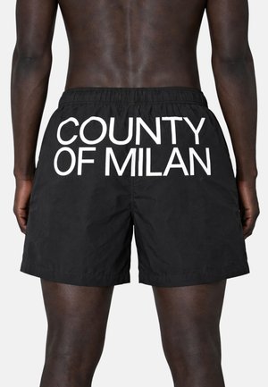 Pantaloncini neri con scritta bianca "COUNTY OF MILAN" sul retro, indossati da una persona senza maglietta vista da dietro, vista a metà coscia.