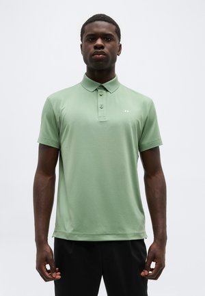 Homme portant un polo à manches courtes vert clair avec un petit logo blanc sur le côté gauche de la poitrine, debout devant un fond uni.