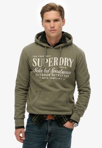 Sudadera verde oliva con bolsillo frontal y capucha con cordón, con texto impreso en blanco. Superpuesta sobre una camisa de cuadros en negro y verde.