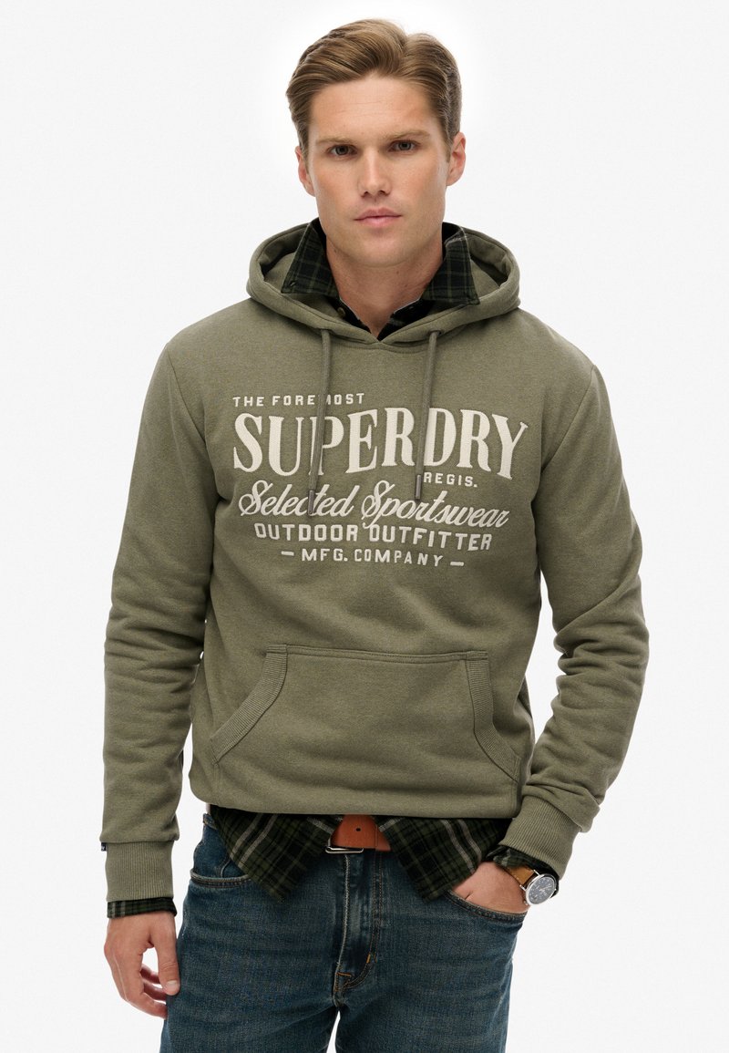 Sudadera verde oliva con bolsillo frontal y capucha con cordón, con texto impreso en blanco. Superpuesta sobre una camisa de cuadros en negro y verde.